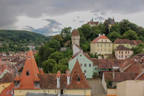 Landscape view from the Sighisoara Clock Tower, looking over the red-tiled rooftops and pastel-colored buildings of the medieval town. The lush green hills and distant landscape provide a scenic backdrop, encapsulating the charm of this Transylvanian village.