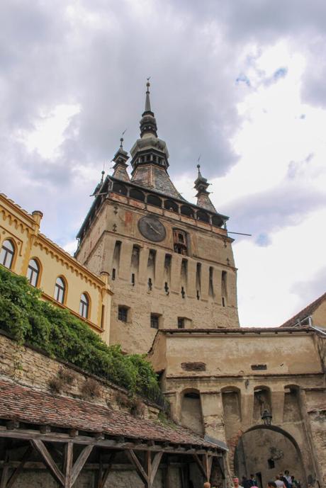 A close-up of the clock tower in Sighișoara, Romania. The medieval tower has a large clock face and ornate spires, seen against a partly cloudy sky, with nearby yellow and beige buildings.