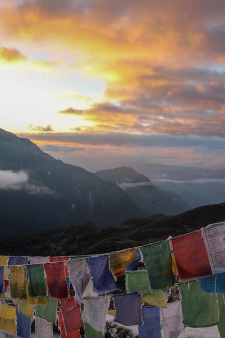 A stunning sunset with a vibrant orange and pink sky illuminating the mountains in the background, as prayer flags flutter in the foreground. The scene captures the serenity of the mountain landscape at dusk.