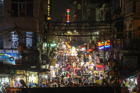 A vibrant, crowded street market at night in Delhi, packed with people and rickshaws. Neon signs for hotels and shops light up the scene, while tangled electrical wires crisscross overhead.