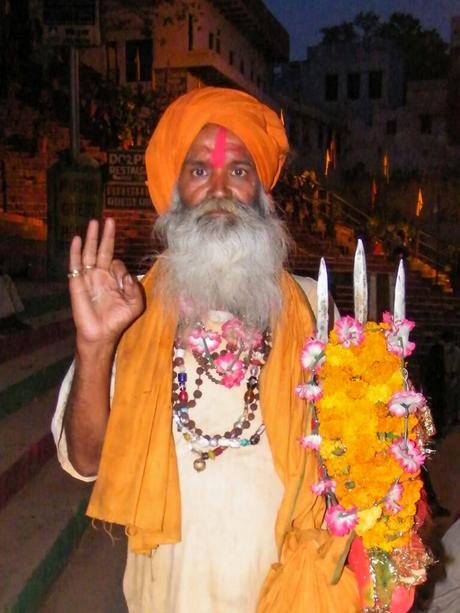 A man with a long white beard and orange turban, wearing beaded necklaces and holding an orange flower garland. He stands against a backdrop of steps at dusk, offering a gesture with his right hand.