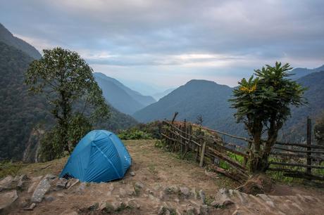 A blue tent pitched on a flat, grassy spot in the mountains, overlooking a vast, misty valley. The mountains stretch out under a cloudy sky, creating a peaceful, isolated camping scene.