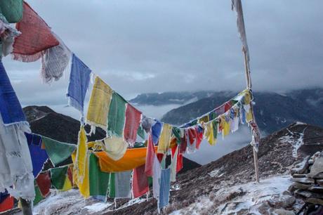 A line of colorful prayer flags strung along a snowy mountain ridge, with mist and clouds hovering over distant mountains. The scene conveys a sense of tranquility and devotion.