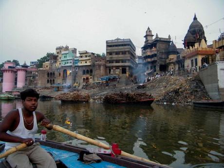 A boatman rows on the Ganges River near a burning ghat, with ancient structures and smoke rising from funeral pyres on the riverbank.