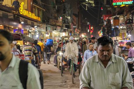 People walking and cycling through a busy street at night, with signs for hotels and shops glowing in the background. Rickshaws and motorbikes move through the crowd.