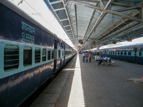 A train station platform in India with a long blue sleeper train, labeled in both English and Hindi, and passengers waiting on benches.