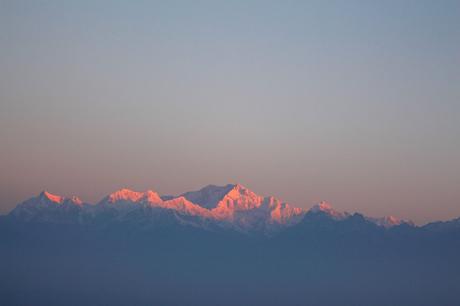 The peaks of the Himalayas bathed in soft pink light as the sun sets or rises, with a hazy sky in the background.