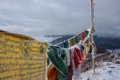 Frost-covered prayer flags fluttering in the wind, against a cold, cloudy mountainous backdrop. The tattered flags display traditional Tibetan symbols and writings, evoking a spiritual atmosphere.