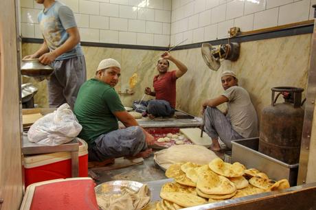 Three men working in a traditional tandoor kitchen, sitting near a clay oven. Freshly baked naan bread is piled in the foreground, while one chef holds a skewer with dough ready to be baked.