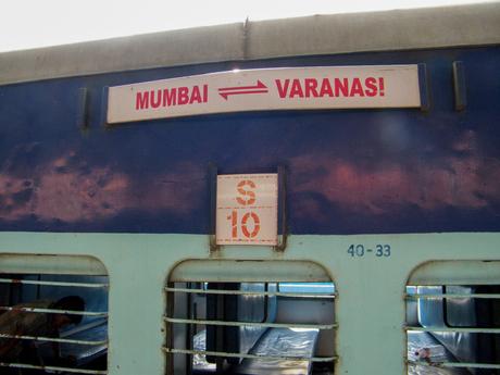 A sign on a blue train coach indicating a route from Mumbai to Varanasi, with an open window revealing a passenger inside.