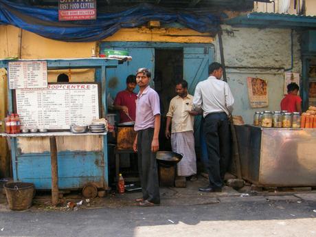 A busy street food stall with a handwritten menu on a board. The vendor stands in front of a rustic blue wooden counter, while others cook behind him, preparing local dishes.