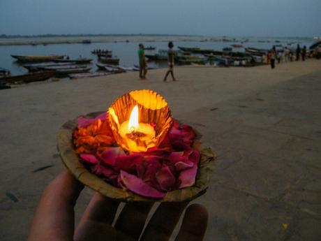 A hand holds a floating flower garland and lit candle, ready to be placed in the Ganges River. Boats and distant figures can be seen along the shore.