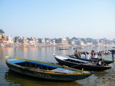 Boats filled with people floating on the Ganges River in Varanasi, with a backdrop of colorful buildings and crowds of people along the ghats.