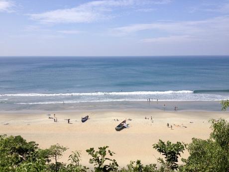 A panoramic view of a serene beach at Varkala with soft sand and gentle waves, scattered with a few people, boats, and beachgoers under a clear blue sky.