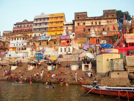 A colorful scene of locals bathing in the Ganges River along a ghat with steps leading up to vibrant, worn-out buildings under the early morning light.