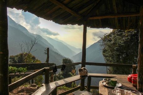 A small shelter overlooking a beautiful mountain valley with layers of forest-covered hills. A cat rests on a wooden bench, and a table with cups and dishes sits in the foreground under the rustic roof.