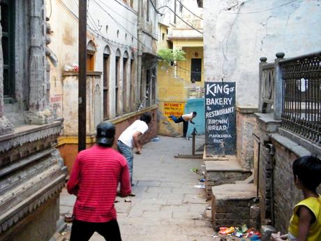 Boys playing cricket in a narrow alley surrounded by old, worn-out buildings. A sign on the side advertises a bakery and restaurant.