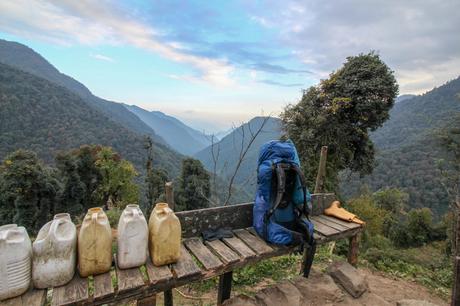 A scenic view of an Osprey backpack resting on a bench in a mountainous landscape, with jugs of water lined up next to it. The lush green valley and misty mountains stretch into the distance under a partly cloudy sky.