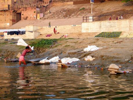 A man washing clothes by the riverbank, slapping wet fabric against rocks, with other laundry laid out to dry on the steps behind him.