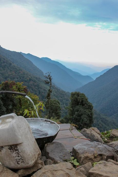 A close-up of a water source in the mountains, with a white container and a flowing spout of water. The misty blue mountains in the background enhance the serenity of the scene.