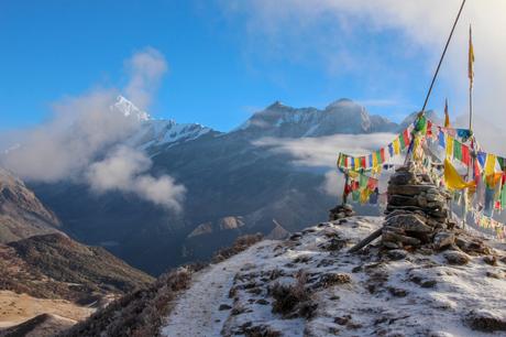 A vibrant, snow-covered mountain peak with colorful prayer flags blowing in the wind at the summit. The clear blue sky and misty clouds add a sense of elevation and awe.