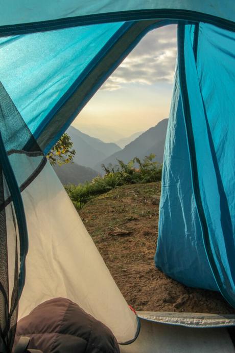 A view from inside a blue tent looking out toward the misty mountains in the early morning light. The tent flap frames the serene landscape, with gentle sunlight filtering through the clouds.