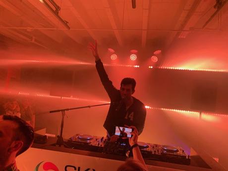 The DJ Mark Ronson raises his hand while playing music in a dimly lit, red-hued venue. A person in the foreground captures the moment on their phone.