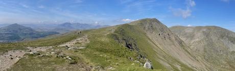 A sweeping panoramic view of a ridgeline in the Lake District, with rocky hills leading to distant peaks. The blue sky stretches across the vast landscape.