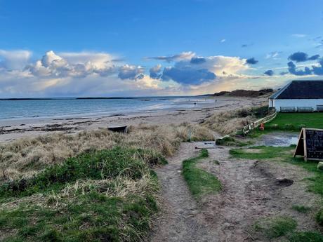 The view captures a wide, sandy beach with waves gently lapping at the shore. In the background, the silhouette of a distant castle ruin stands against a colorful sky.