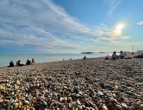 A rocky beach in Sussex under a sunny sky, where small groups of people sit near the shore. In the distance, a pier juts out into the calm sea.