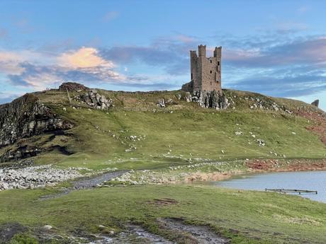 A solitary castle tower stands on a grassy hill surrounded by rocky outcrops. The sky is lightly clouded, with soft evening light illuminating the scene.