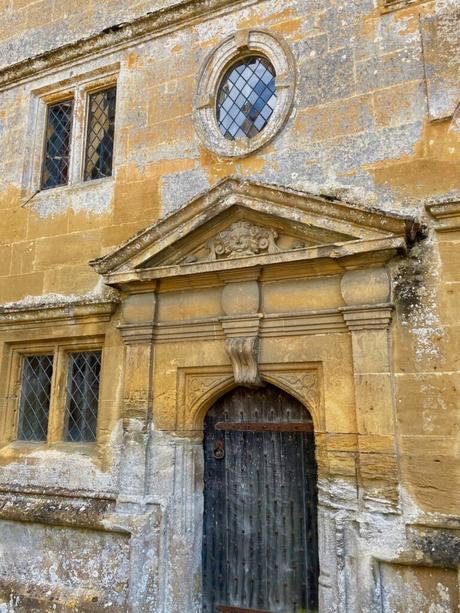 A weathered stone building with a detailed, arched doorway, flanked by small windows with diamond-paned glass. The circular window above adds to the charm of the historic architecture.