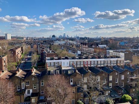 A wide view of North London’s residential rooftops stretching toward the city skyline. The sky is bright blue, with scattered clouds and clear visibility of famous landmarks in the distance.