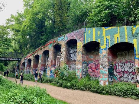 A line of brick arches, heavily decorated with colorful graffiti, runs along a dirt path in a park. People walk along the trail beneath the vibrant street art.