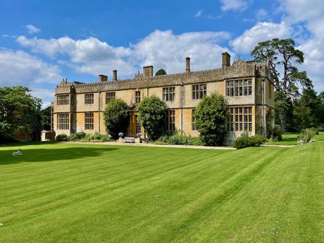 A grand stone manor house with large windows and a meticulously maintained lawn. The sky is clear, and trees surround the stately home.