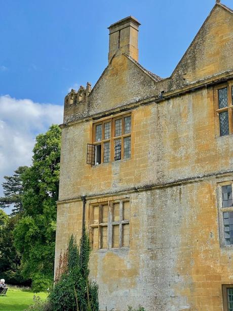 The side of an aged stone manor with tall windows and a prominent chimney. The building is surrounded by lush greenery under a bright blue sky.