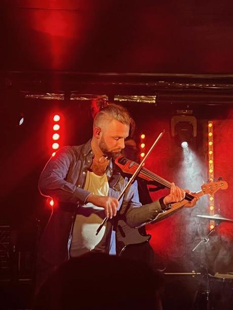 A close-up of Alex Tiffany performing on stage playing the electric violin under warm red and yellow stage lights. He is deeply focused, creating an intimate concert moment.