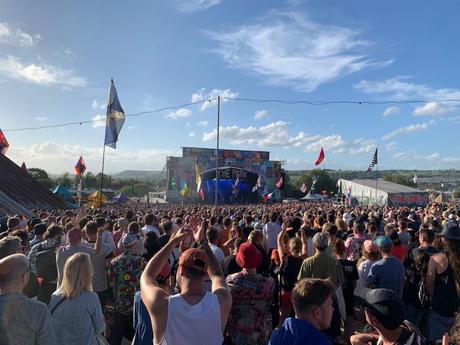 A massive crowd gathers at the outdoor Glastonbury music festival, facing a stage adorned with colorful flags. The sky is clear, with vibrant decorations and festival energy filling the atmosphere.