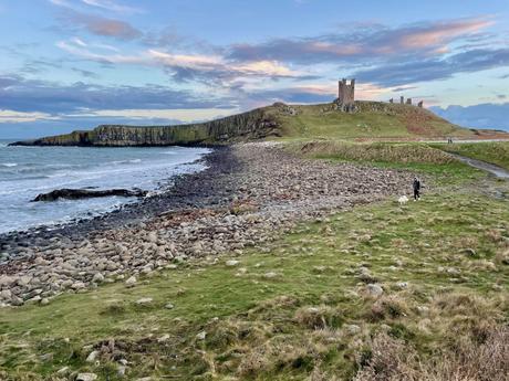 A rocky shoreline leads to the ruins of a castle perched on a grassy hill. The waves of the sea gently lap against the stony beach under a colorful sky.