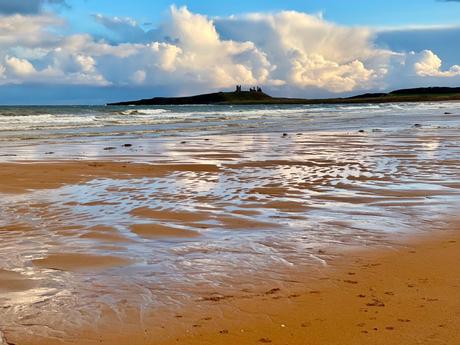 A picturesque beach scene, with shallow waves rolling onto the wet sand. The sky is filled with large, dramatic clouds, and a distant island with ruins adds a historic element to the view.