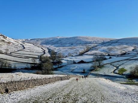 Rolling hills in the Yorkshire Dales covered in a light dusting of snow are divided by winding stone walls. A few people are seen walking in the distance, enjoying the quiet winter scenery.