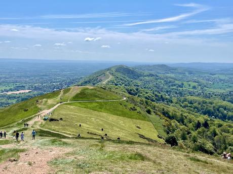 A scenic ridgeline with a well-trodden hiking trail winding through green hills in Worcestershire. Distant hills and valleys stretch out under a bright blue sky.