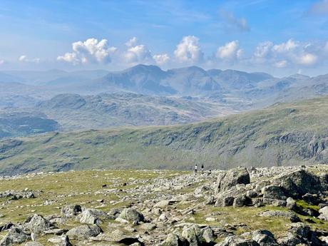 View from the top of the Old Man of Coniston with rugged, rocky terrain, and distant mountains fading into the horizon under a blue sky with soft clouds.