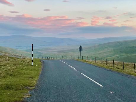 A quiet road winds through a hilly landscape in the North Pennines, with a dramatic pink and orange sky from the setting sun. The landscape is serene and expansive.