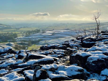 Snow-dusted stone walls and patches of grass stretch across a rural, hilly landscape. In the foreground there is the unusual limestone pavement at the top of Malham Cove in Northern England. The distant hills are blanketed with light snow under a soft winter sky.