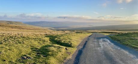 A narrow road cuts through an expansive, grassy landscape under a setting sun. Rolling hills stretch into the distance, bathed in soft evening light.