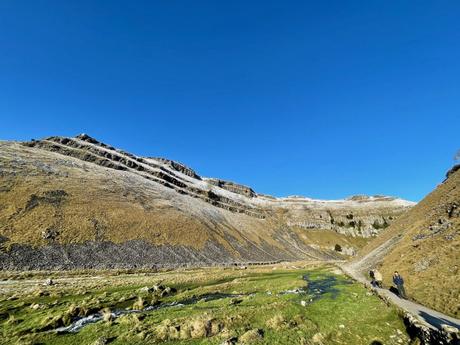 A steep hill with rugged rock formations at Gordale Scar under a bright blue sky. A winding path leads through the valley below, with a few hikers in the distance.