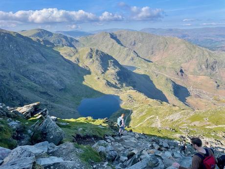 Hikers navigate a steep mountain path, overlooking a valley with lakes nestled between tall, rocky mountains. The sun casts shadows over parts of the landscape.