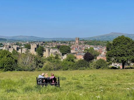 A group of three people sits on a bench, gazing at the town of Ludlow with its large church and castle ruins. The lush countryside stretches out in the distance under a clear sky.