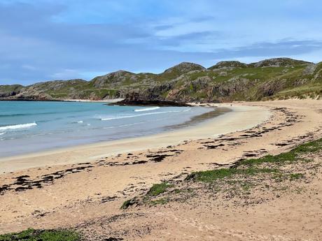 A wide, curving sandy beach with scattered seaweed, stretching into the distance and framed by low, grassy hills.